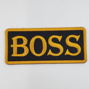 May include: Black and gold embroidered patch with the word "BOSS" in a bold, stylized font.