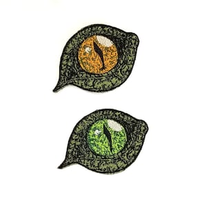 May include: Two embroidered patches depicting stylized green cat eyes with orange and green pupils.