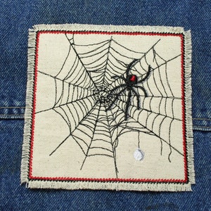 Puede incluir: Un parche de tela blanca bordado con una araña negra con una mancha roja en la espalda. La araña está sentada en el centro de una telaraña negra. El parche tiene un borde rojo y negro.