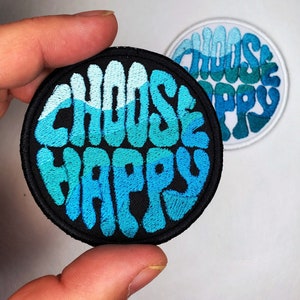 Puede incluir: Un parche redondo negro con las palabras "Choose Happy" bordadas en azul y turquesa. Las letras son estilizadas y tienen un diseño ondulado, como el océano.