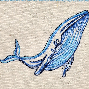 Op de afbeelding: Een blauwe walvis geborduurd op een beige vierkant van stof met een blauwe en witte rand.