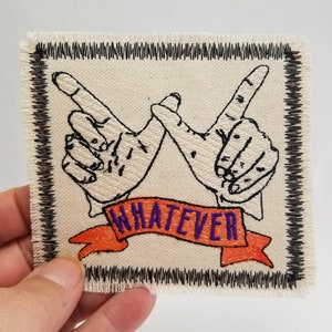 Pode incluir: Um remendo de tecido branco com costuras pretas ao redor das bordas. O remendo apresenta duas mãos fazendo o gesto de "tanto faz" com uma faixa de fita laranja que diz "WHATEVER" em preto com letras roxas.