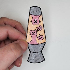 May include: An embroidered iron-on patch of a lava lamp with three pink cats inside. The lava lamp is yellow and has a gray base.