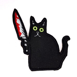 May include: Black cat with green eyes holding a red and white knife. The cat is sitting on a white background.