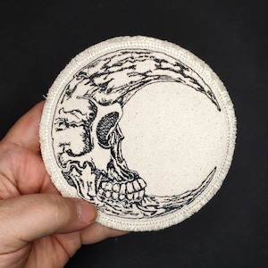 May include: Round patch featuring a black and white illustration of a crescent moon with a skull design. The patch has a cream-colored background and a stitched border. The design is detailed and gothic in style.