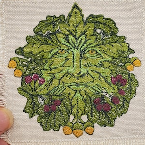 May include: A green man's face embroidered on a beige fabric patch with a frayed edge. The face is surrounded by green leaves, red berries, and yellow acorns.