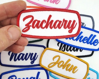 Op de afbeelding: Geborduurde naamlabels in verschillende kleuren. De labels hebben een witte achtergrond met een gekleurde rand en tekst. Namen zijn onder meer "Zachary", "Pink", "Michelle", "John", "Royal", "Navy" en "Jose".