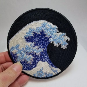 May include: A round embroidered patch featuring a blue and white wave design on a black background. The wave is a stylised depiction of the Great Wave off Kanagawa, a famous Japanese woodblock print.