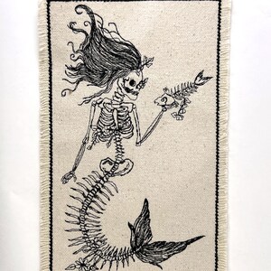 May include: A black and white embroidered patch of a skeleton mermaid holding a fish. The patch is on a beige fabric background with a frayed edge.