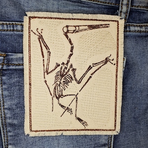 May include: A brown embroidered patch of a dinosaur skeleton on a beige background. The dinosaur is in a flying pose with its wings outstretched.