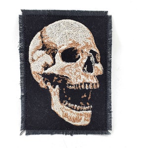 May include: A black fabric patch with a white embroidered skull with brown details. The skull is facing right and has an open mouth.
