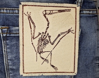 May include: A brown embroidered patch of a dinosaur skeleton on a beige background. The dinosaur is in a flying pose with its wings outstretched.
