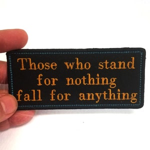 Peut inclure: Écusson brodé noir avec des coutures blanches autour des bords. L'écusson présente le texte "Those who stand for nothing fall for anything" en fil orange.