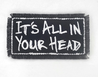 Puede incluir: Parche bordado negro con costuras blancas. El parche dice "It's All in Your Head".