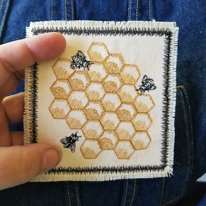 May include: A white fabric patch with a black border, featuring an embroidered honeycomb design in gold and three black bees.