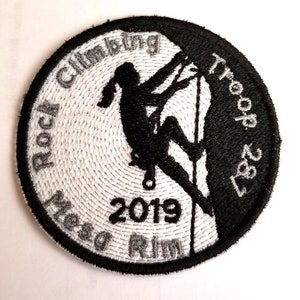 May include: Black and white embroidered patch with a silhouette of a person rock climbing. The text "Rock Climbing Troop 327" is on the right side of the patch. The text "Mesa Rim 2019" is on the left side of the patch.