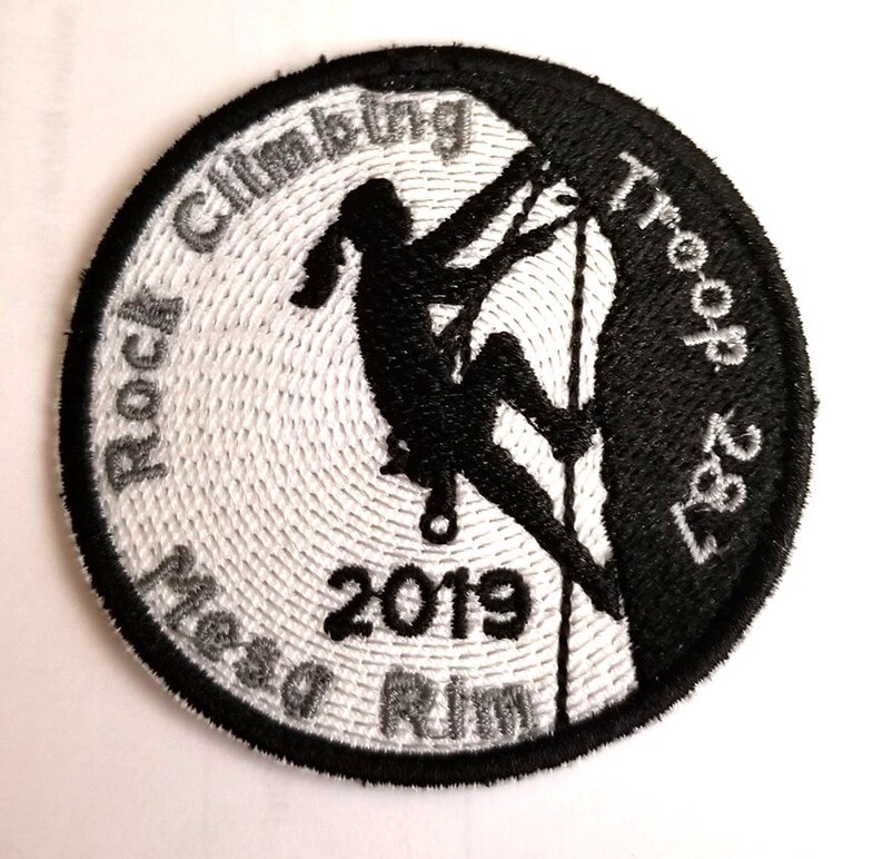 May include: Black and white embroidered patch with a silhouette of a person rock climbing. The text "Rock Climbing Troop 327" is on the right side of the patch. The text "Mesa Rim 2019" is on the left side of the patch.