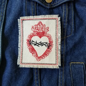 May include: A red embroidered heart with a crown of thorns and flames, on a white fabric patch with frayed edges. The patch is sewn onto a blue denim jacket.