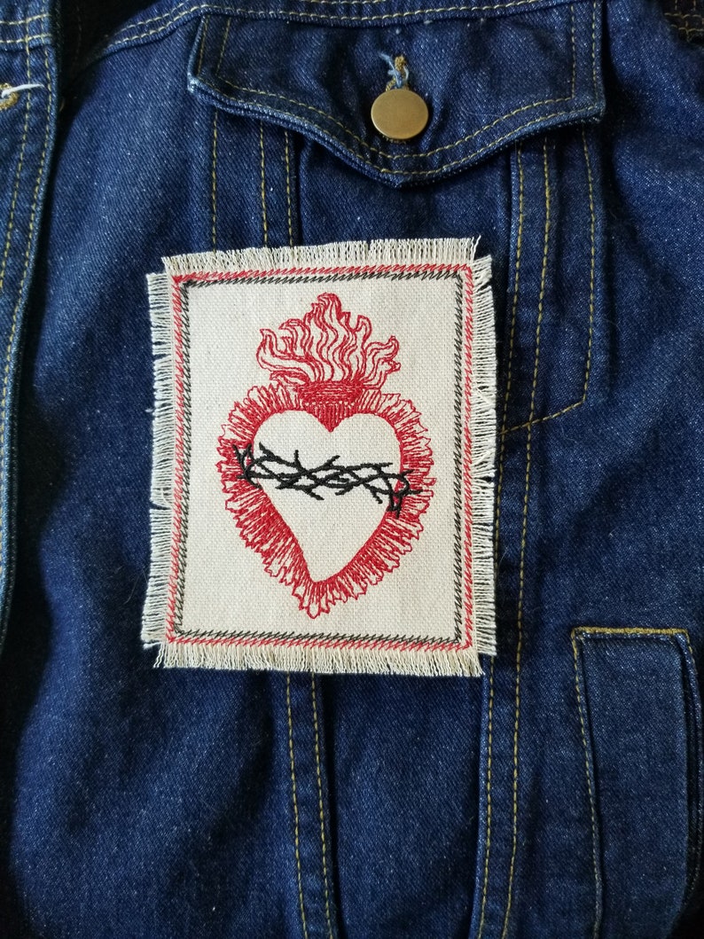 May include: A red embroidered heart with a crown of thorns and flames, on a white fabric patch with frayed edges. The patch is sewn onto a blue denim jacket.