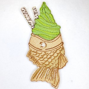 May include: A green matcha soft serve ice cream in a crispy taiyaki fish-shaped waffle cone with two brown and white striped straws.