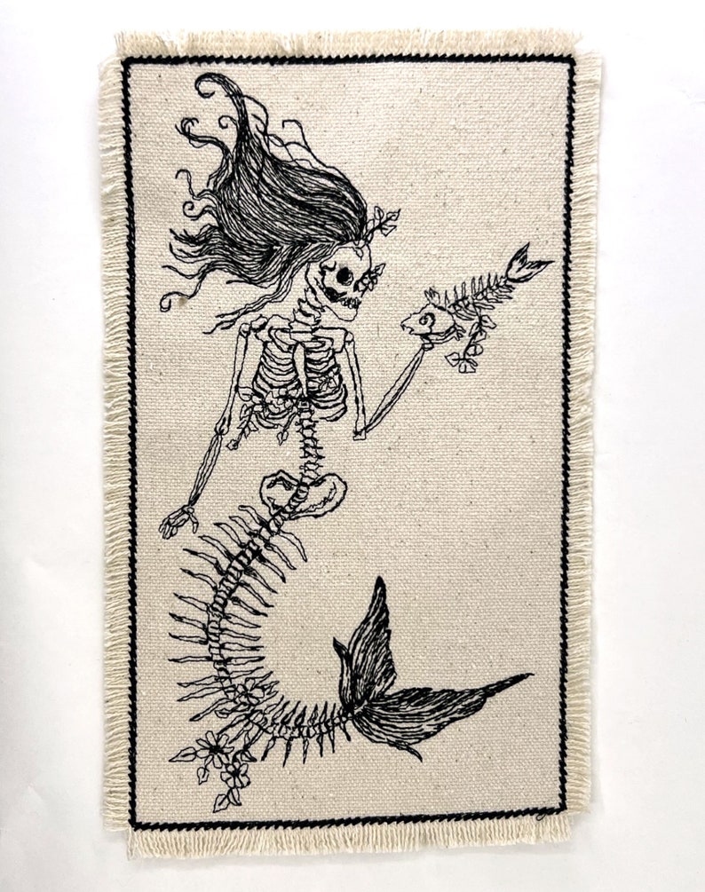 May include: A black and white embroidered patch of a skeleton mermaid holding a fish. The patch is on a beige fabric background with a frayed edge.