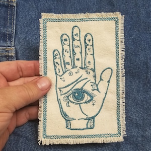 May include: A blue embroidered patch featuring a hand with an eye in the palm. The hand has lines and symbols on the fingers and palm. The patch has a frayed edge.