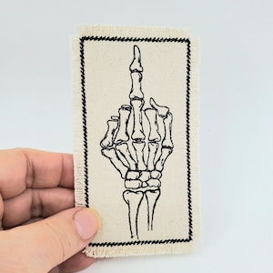 May include: A rectangular patch with a black stitched border and a black embroidered skeleton hand giving the middle finger. The patch is made of a beige fabric.