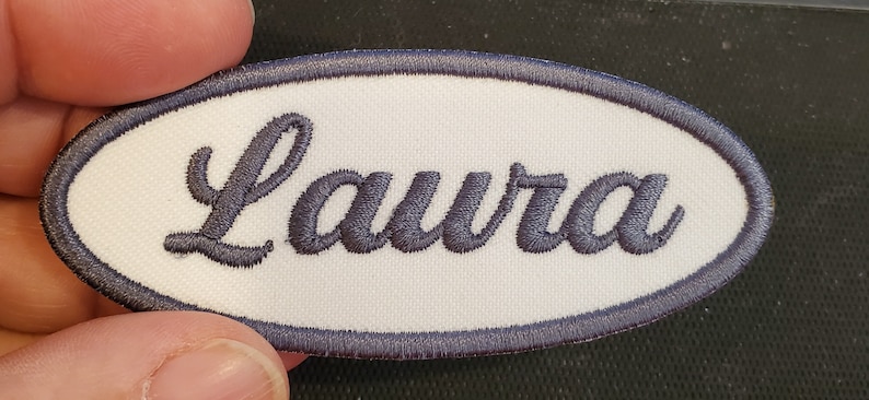 May include: White oval-shaped fabric patch with a dark blue border and the name "Laura" embroidered in a cursive font.