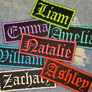 May include: A set of nine iron-on patches featuring different names in a variety of colours. The names are Liam, Emma, Amelia, Natalie, William, Ashley, Zachary, and two others. The patches are displayed on a blue denim background.