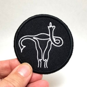 May include: A black embroidered patch with a white outline of a uterus with a middle finger raised in the air.