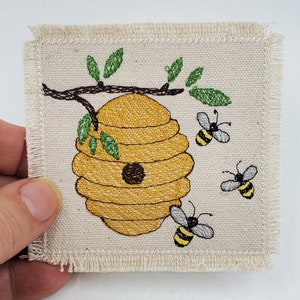 Pode incluir: Um quadrado de tecido bege com uma colmeia bordada em amarelo e marrom com três abelhas voando ao redor. A colmeia está em um galho com folhas verdes.