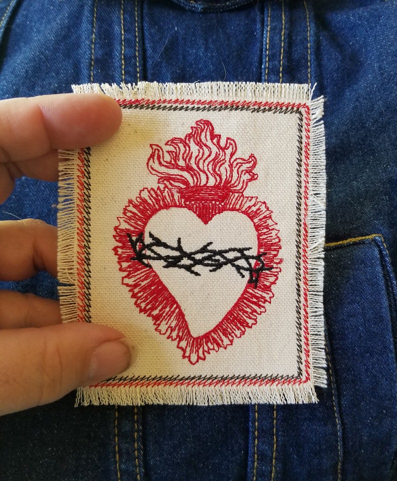 May include: A red embroidered heart with a crown of thorns and flames, on a white fabric patch with a frayed edge.