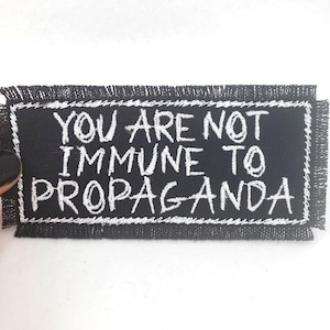 Può includere: Toppa ricamata nera con cuciture bianche. La toppa riporta la scritta "You are not immune to propaganda."