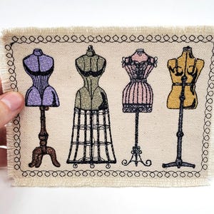 May include: A fabric patch featuring an embroidered design of four vintage dress forms on stands. The dress forms are in various colours, including purple, green, pink, and yellow.