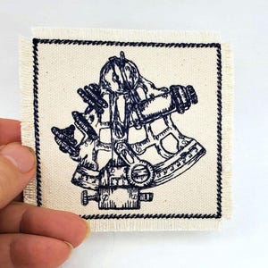 May include: A navy blue embroidered patch depicting a sextant, a nautical instrument used for measuring angles, on a white canvas background.