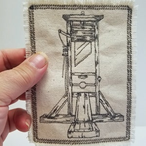 May include: A black and white illustration of a guillotine on a beige fabric patch with a brown border.