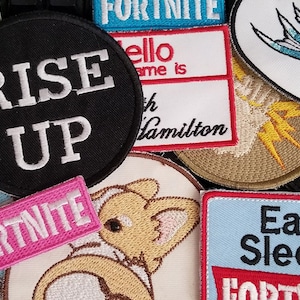 May include: A collection of embroidered patches featuring various designs, including "Rise Up", "Fortnite", "Eat Sleep", "Bowie", and a cartoon fox. The patches are colorful and feature a variety of fonts and styles.