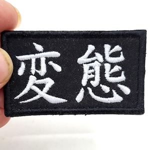 Peut inclure: Écusson rectangulaire noir avec des caractères japonais brodés en blanc. L'écusson est tenu par une main sur fond blanc. Le texte est dans une police stylisée.