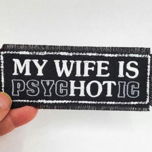 May include: Black rectangular patch with white embroidered text that reads "MY WIFE IS PSYCHOTIC". The patch has a white border and frayed edges. The patch is held by a hand against a white background.