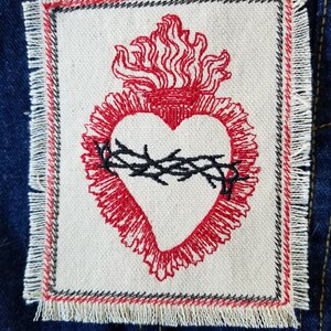 May include: A red embroidered heart with a crown of thorns and flames, on a white fabric patch with a frayed edge.