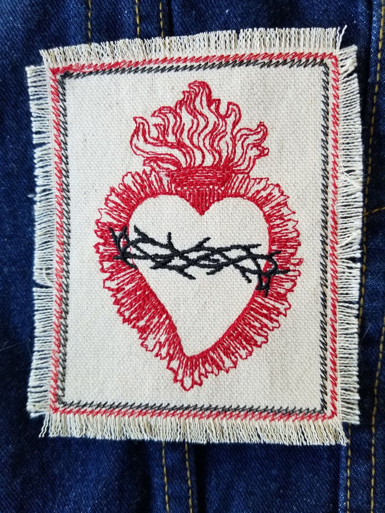 May include: A red embroidered heart with a crown of thorns and flames, on a white fabric patch with a frayed edge.