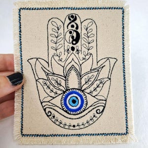Könnte beinhalten: Ein weißer Canvas-Patch mit einer schwarzen gestickten Hamsa-Hand mit einem blauen bösen Auge in der Mitte. Die Hamsa-Hand hat ein Yin-Yang-Symbol in der Handfläche und florale Designs um die Finger.