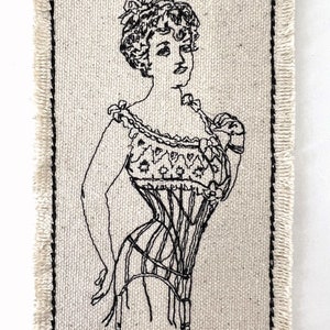 May include: Black and white embroidered image of a woman wearing a corset on a beige fabric background. The fabric has a frayed edge.