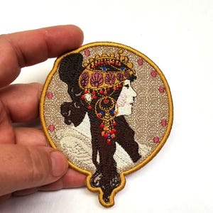 Op de afbeelding: Geborduurde strijkpatch met een vrouw in profiel die een kroon en sieraden draagt. De patch is rond en heeft een gouden rand.