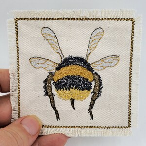 May include: Embroidered bumble bee patch with black and yellow stripes on a white canvas background. The patch has a gold stitched border and frayed edges.