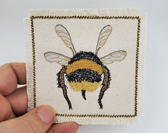 May include: Embroidered bumble bee patch with black and yellow stripes on a white canvas background. The patch has a gold stitched border and frayed edges.