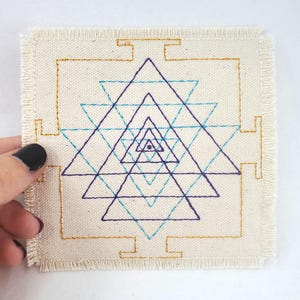May include: A square piece of fabric with a geometric pattern embroidered in blue, teal, and purple thread. The pattern is a series of concentric triangles, with a small circle in the center.