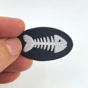以下が含まれることがあります： 白い刺繍の魚の骨格のデザインが施された黒い楕円形のワッペンです