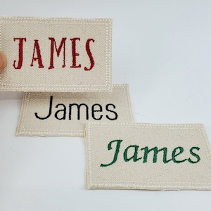 Puede incluir: Tres parches rectangulares de tela beige con el nombre "JAMES" bordado en hilo rojo, negro y verde. Cada parche tiene un borde cosido, y el texto está en una fuente cursiva.
