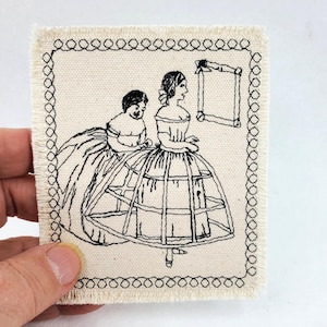 May include: Embroidered patch featuring a black and white illustration of two women in period dresses. The women are standing near a frame. The patch has a cream-colored background with a black stitched border.